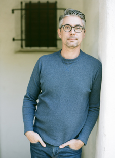 gray-haired man with glasses and dark aqua shirt leaning against a wall