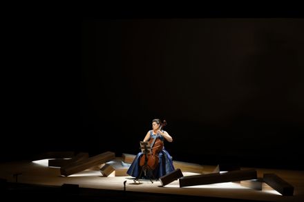 cellist in formal blue gown performs alone on stage amid long rectangular blocks